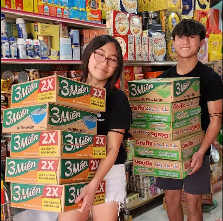 Rilina and Dylan Tran in a store aisle holding stacks of instant noodle cases